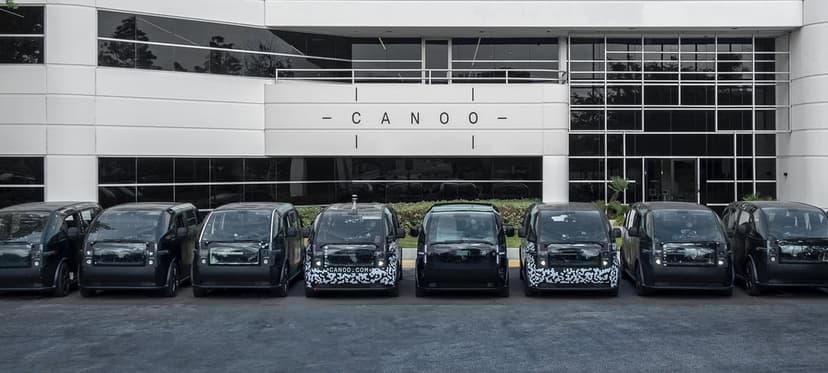 Line of pod-shaped Canoo electric vehicles in black and camouflage parked at a white modern building.
