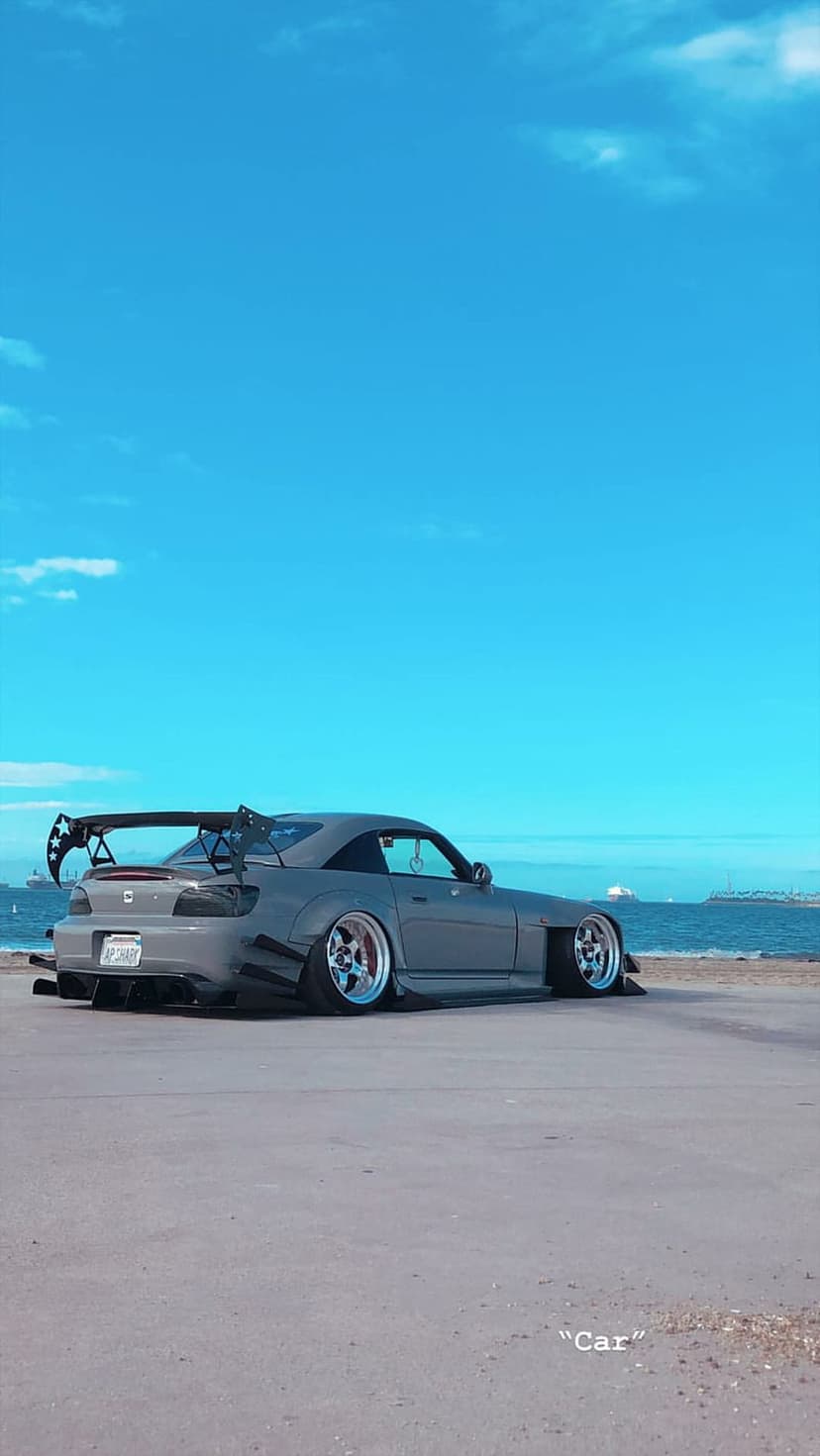 Gray Honda S2000 with a widebody kit and rear spoiler parked on coastal asphalt against a calm blue ocean backdrop.