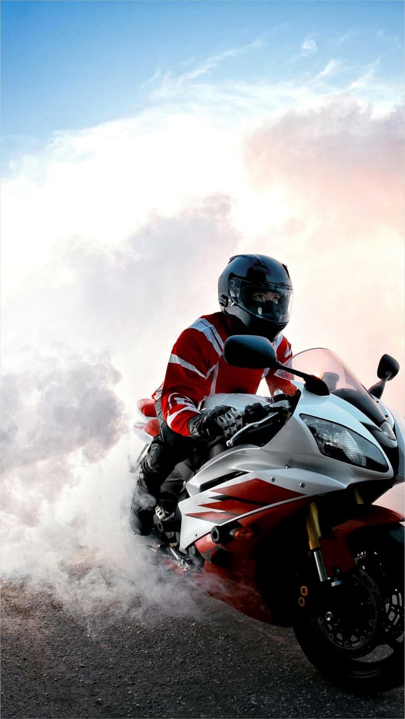 Motorcyclist in red racing leathers performing a rear tire burnout surrounded by thick white smoke at sunset.