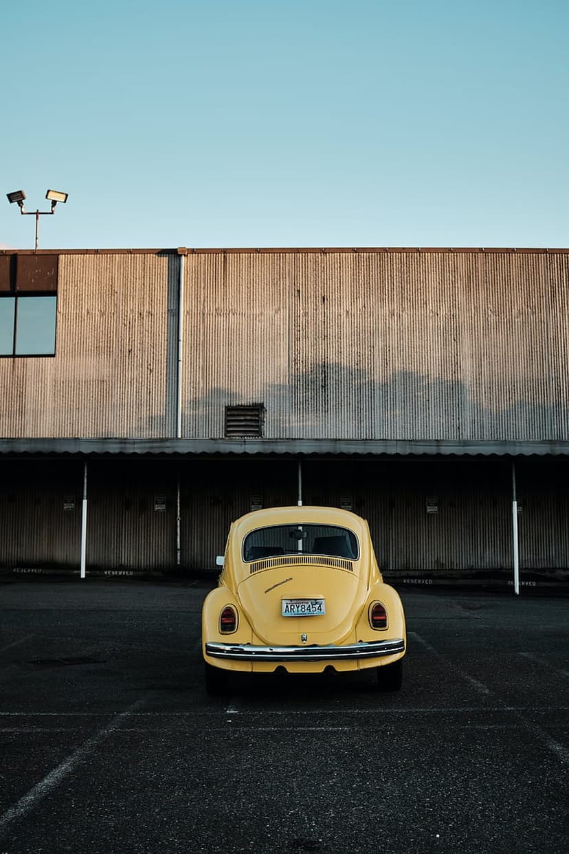 Yellow Volkswagen Beetle rear view parked at an industrial site under a blue sky with long afternoon shadows.