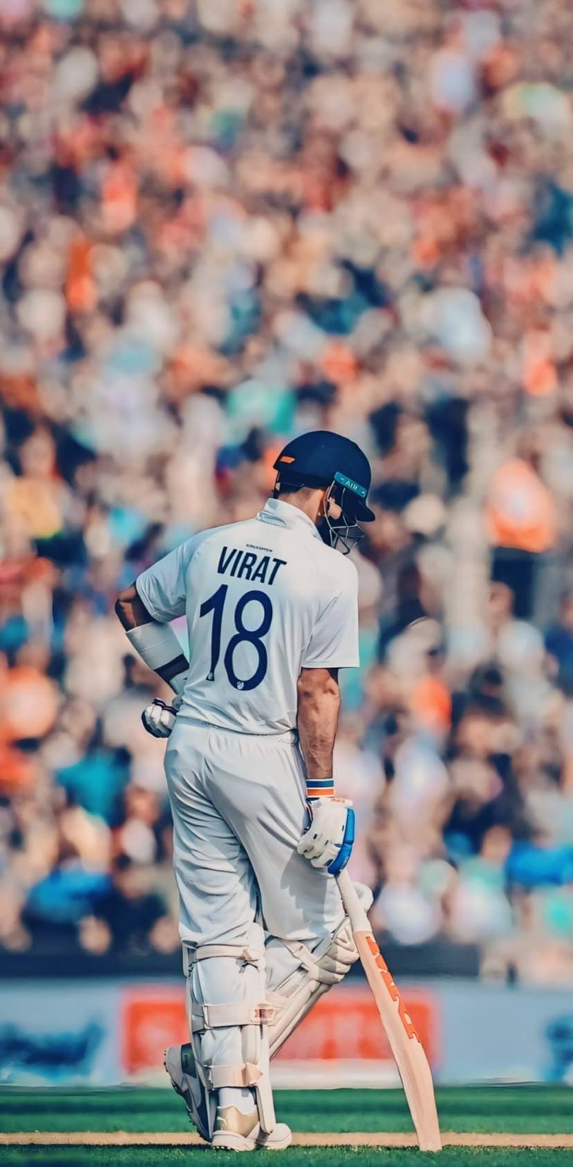 Virat Kohli in a white number 18 jersey holds a cricket bat in a batting stance against a blurred stadium.