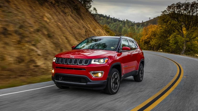 Red Jeep Compass SUV driving through a sharp turn on a winding mountain road with autumn trees and motion blur.
