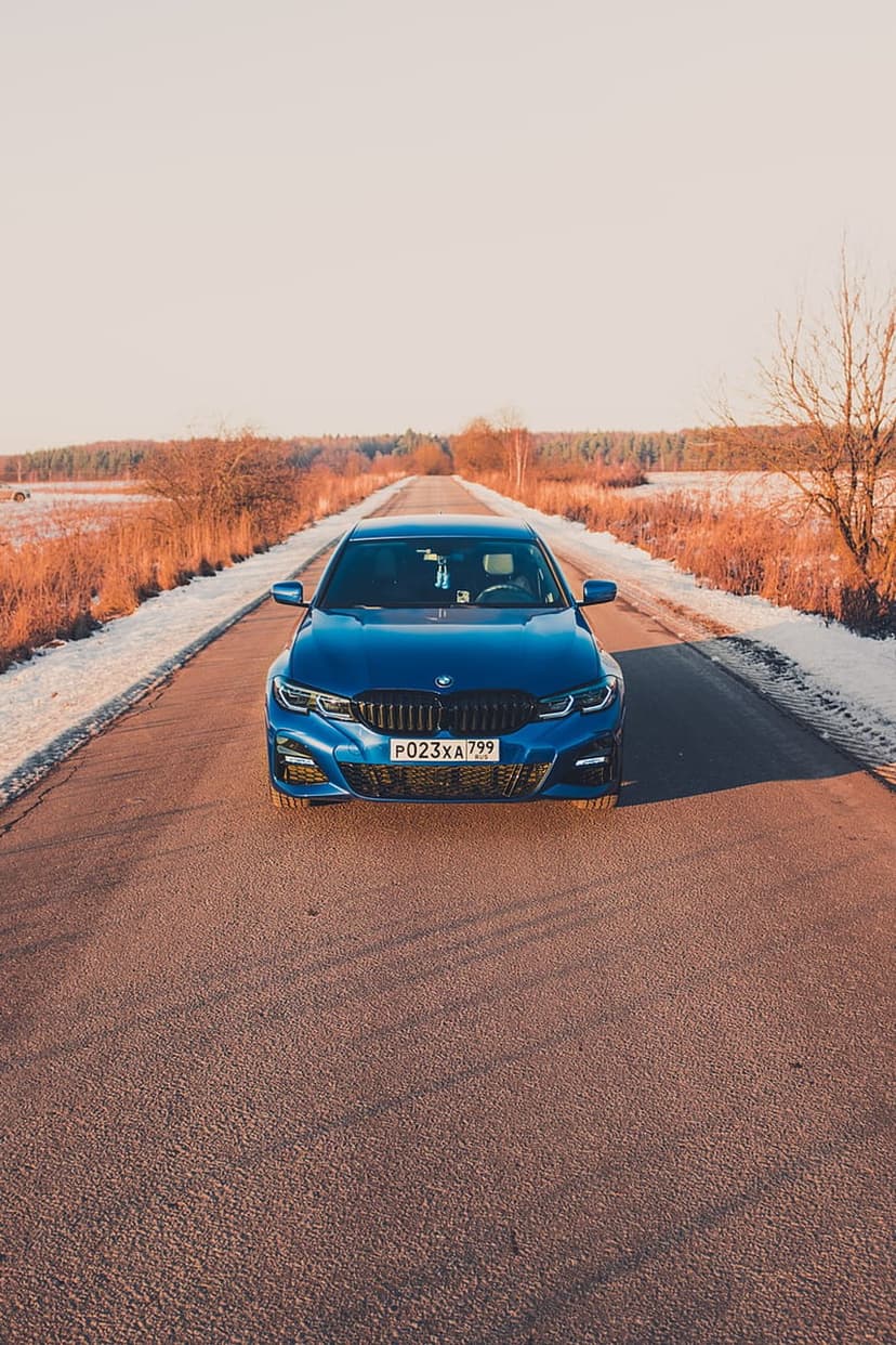 Blue BMW sedan driving on a paved road between snowy banks under warm golden sunlight and a clear winter sky.