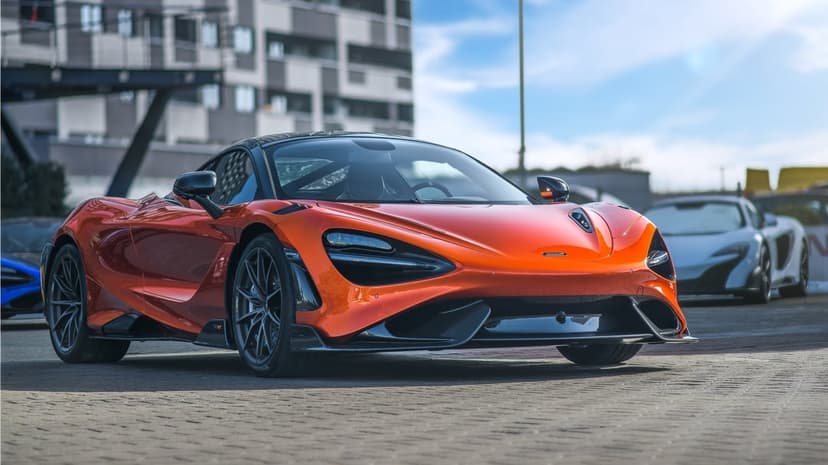 Metallic orange McLaren 765LT supercar parked on pavement with modern buildings and blue sky in the background.