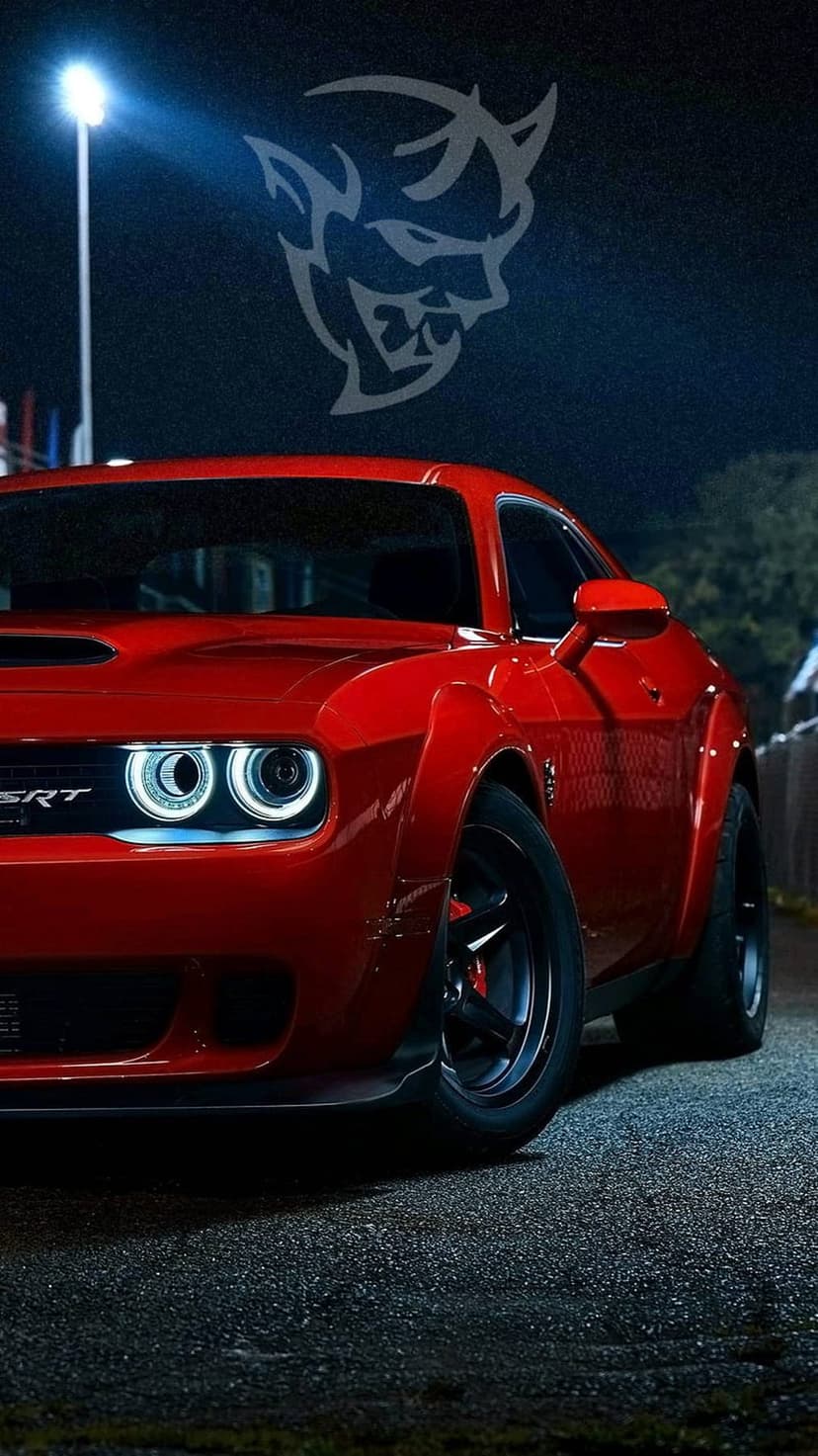 Red Dodge Demon with bright headlights parked on asphalt at night beneath a glowing red devil logo.