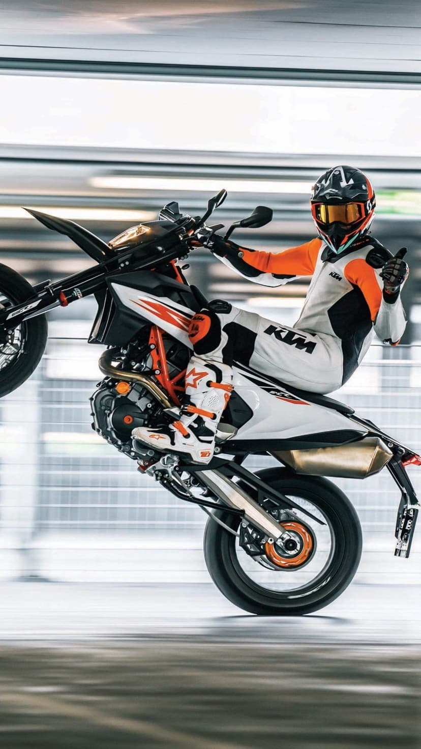 Motorcyclist in racing gear performs a wheelie on an orange KTM bike while giving a thumbs up in an indoor arena.