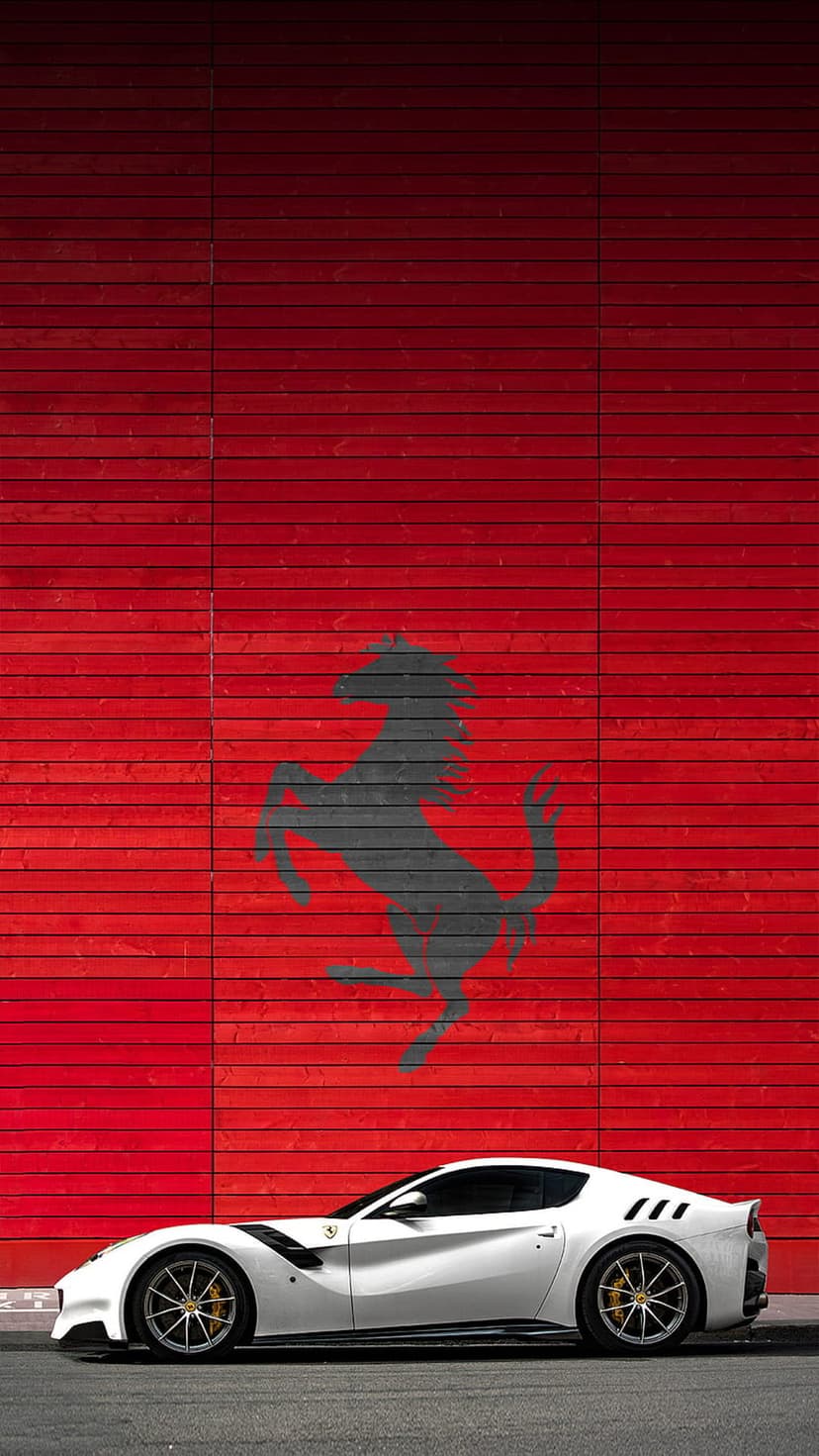 White Ferrari F12berlinetta sports car parked in front of a red wooden wall with a black prancing horse logo.