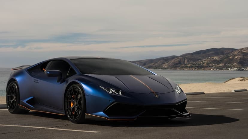 Blue Lamborghini Huracan with orange accents parked on a coastal road against a backdrop of ocean and mountains.