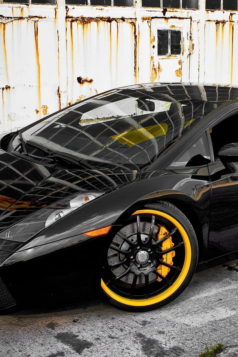 Black Lamborghini with bright yellow rims parked in front of a weathered, rusty white industrial metal wall.
