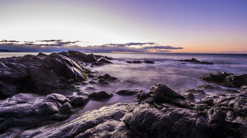 Rugged dark rocks meet misty ocean waves under a vibrant purple and orange twilight sky during high tide.