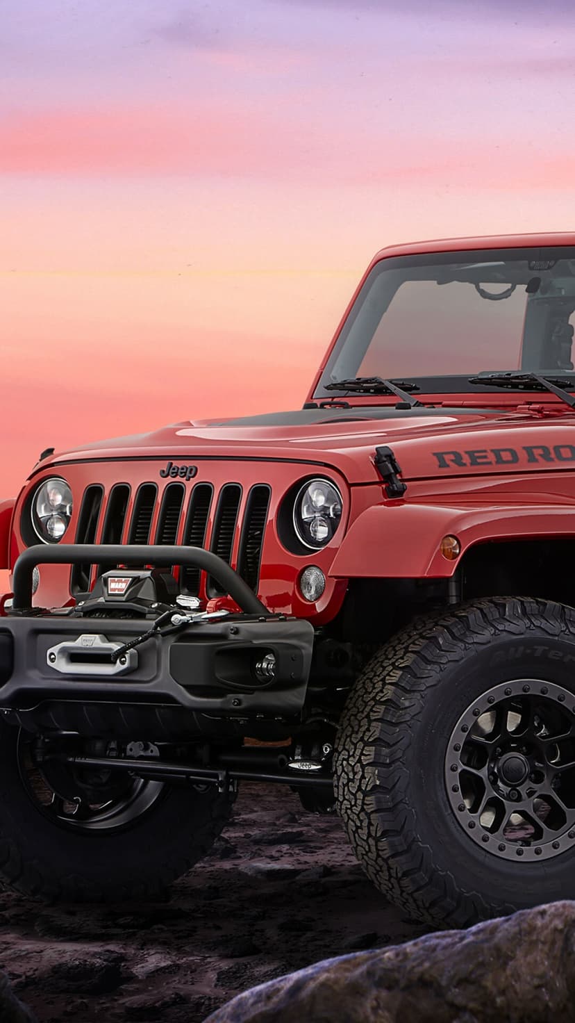 Red Jeep Wrangler with off-road tires and winch parked on rocky ground under a soft pink sunset sky.