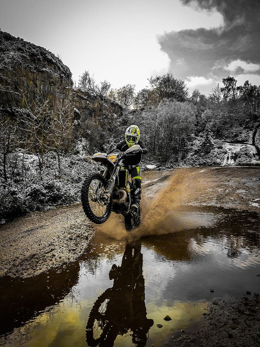 Motocross rider in yellow gear jumping over a muddy water splash amidst snowy rocks and winter forest trees.