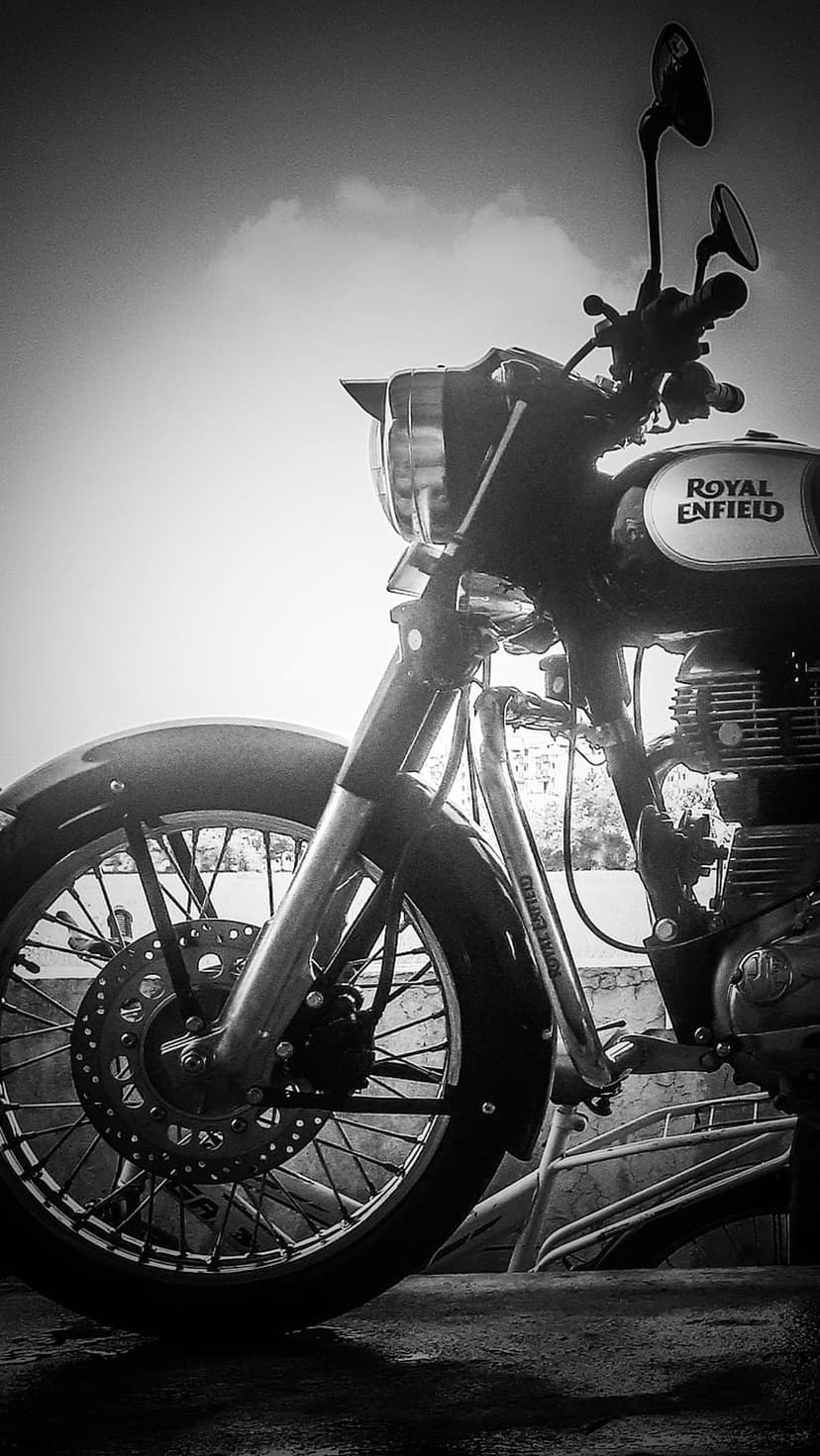 Royal Enfield motorcycle front wheel and fuel tank in black and white against a cloudy sky with engine details.