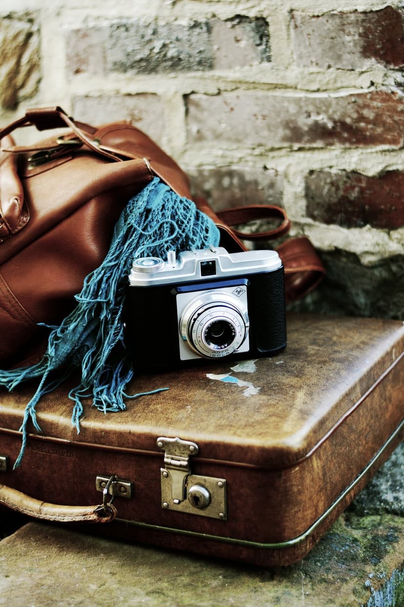 Vintage film camera rests on a worn brown suitcase beside a leather bag against a rustic red brick wall.