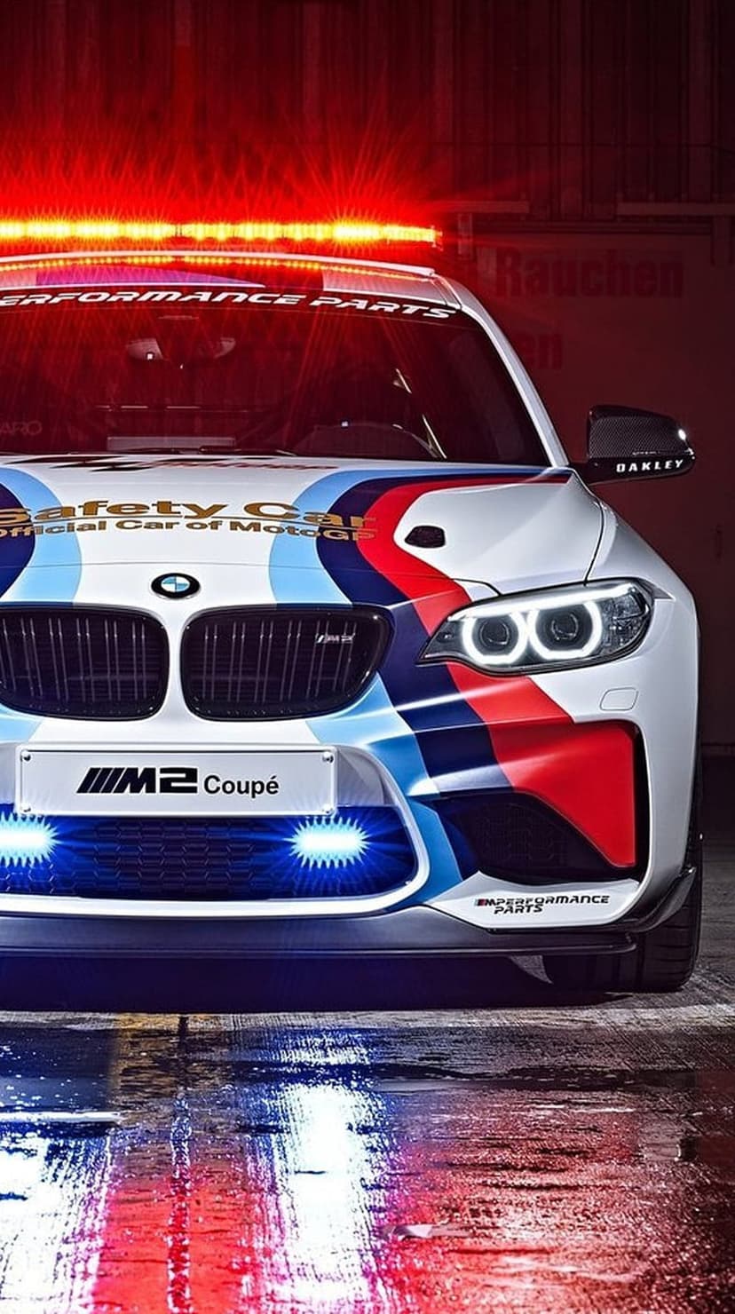 BMW M2 Safety Car with tri-color livery and glowing red light bar reflects on wet asphalt under blue LED lights.