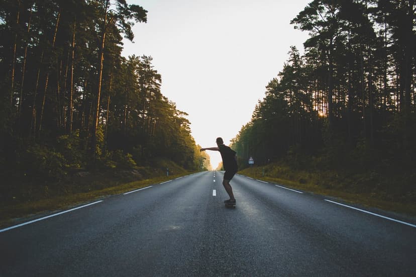 Skateboarder silhouette glides on an asphalt forest road during a warm golden hour sunset with long shadows.