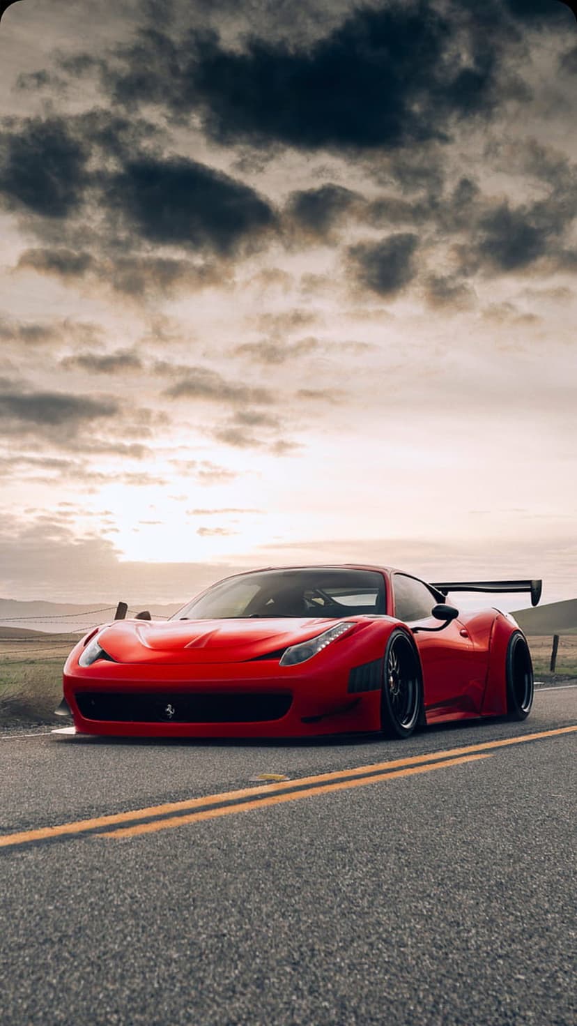 Red Ferrari supercar with a large spoiler parked on a paved road during a dramatic sunset under stormy clouds.