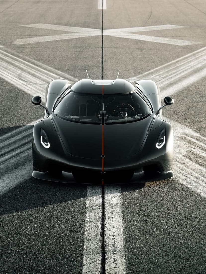 Black Koenigsegg Jesko with an orange racing stripe sits on a racetrack with sharp LED headlights visible.
