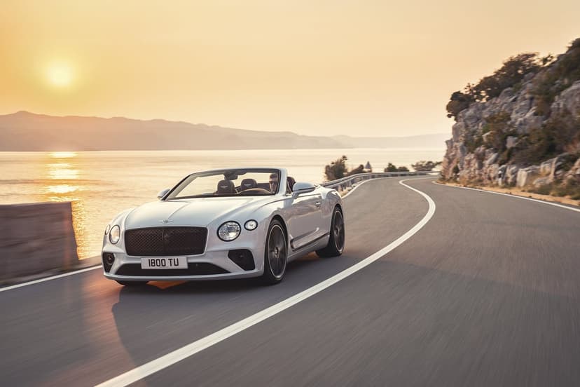 Silver Bentley convertible driving on a winding coastal road next to the ocean at golden hour sunset.
