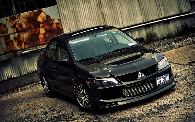 Modified black Mitsubishi Lancer Evolution with a carbon fiber hood parked in front of a rusty industrial metal wall.