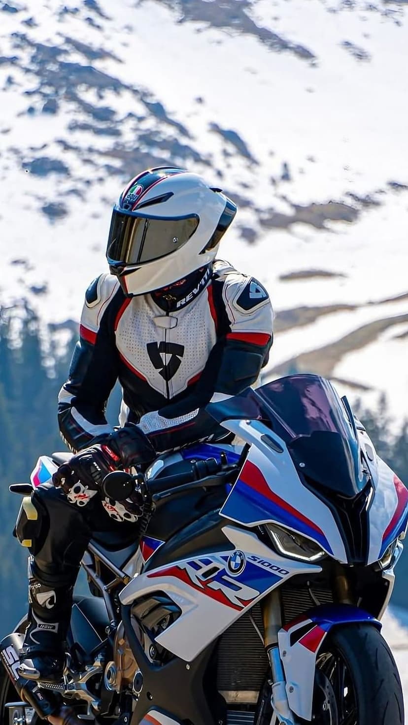 Rider in a white racing suit on a BMW S1000RR motorcycle amidst snow-covered mountains and evergreen trees.
