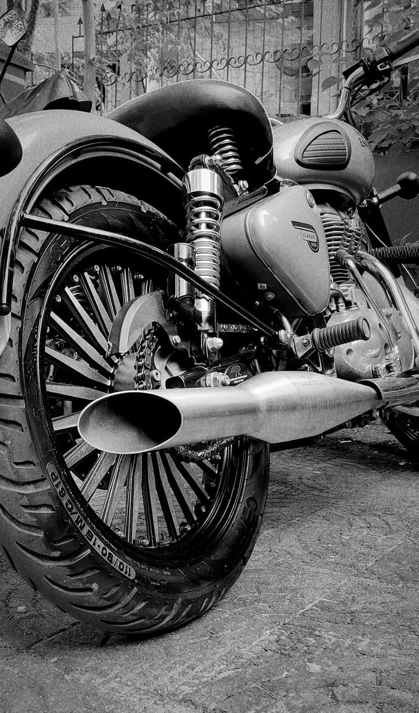 Spoked rear wheel and chrome exhaust system of a vintage motorcycle in a high-contrast monochrome photograph.