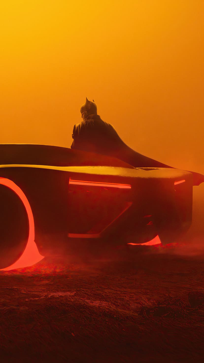 Batman in his silhouette Batmobile cockpit illuminated by red light against a vibrant orange hazy sky background.