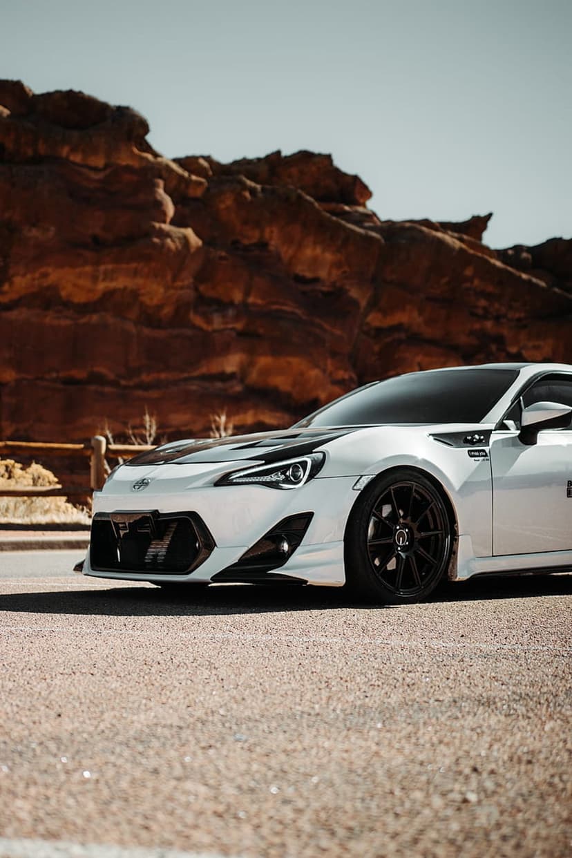 White Toyota GT86 sports car with a black hood on a desert road near massive red rock sandstone formations.