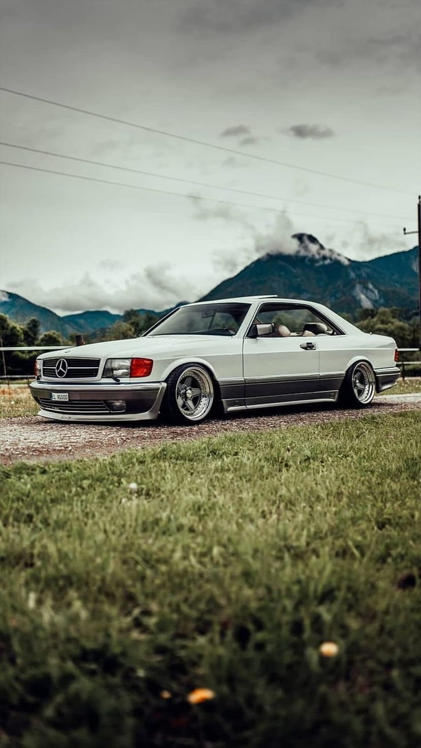 White Mercedes W126 SEC coupe parked on a gravel path with misty mountain peaks and green grass in the background.
