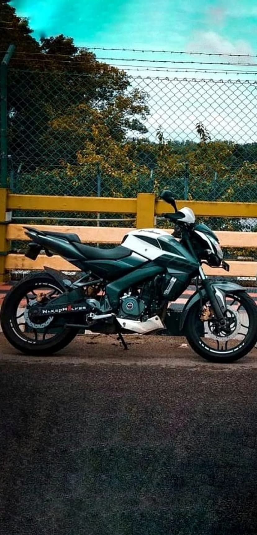 Dark green and white sport motorcycle parked on asphalt with a yellow fence and trees under a clear blue sky.