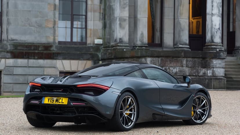 Rear view of a dark grey McLaren 720S supercar on a gravel path in front of a stone building with yellow calipers.