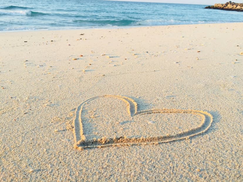 Heart shape etched in golden sand as white sea foam rolls over the shoreline under soft coastal lighting.