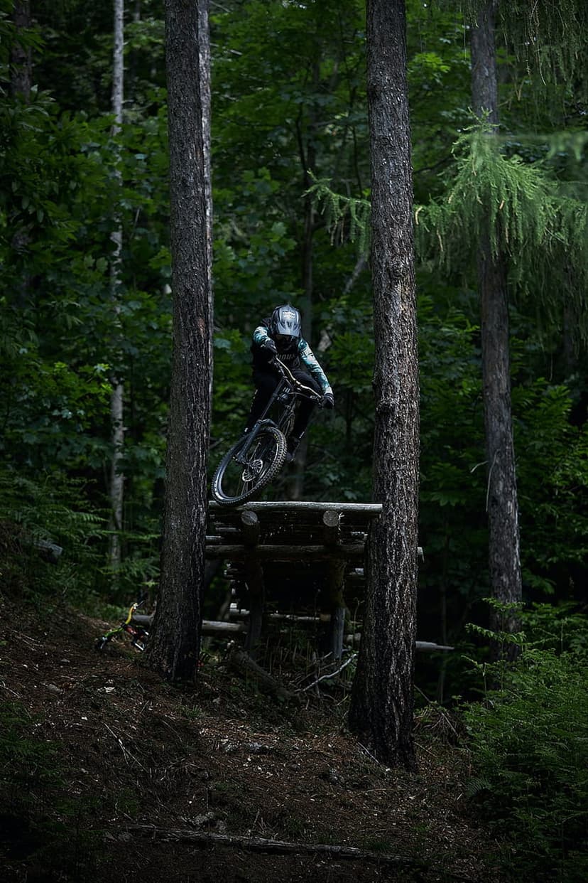 Mountain biker in mid-air jumping a wooden ramp surrounded by dense green trees and sunlit forest foliage.