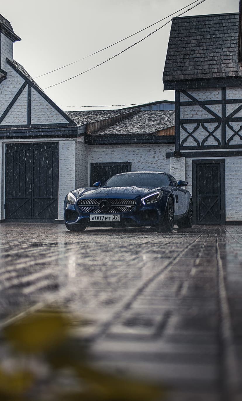 Dark blue Mercedes-AMG GT S parked on wet cobblestones in front of a white and timber Tudor building in the rain.