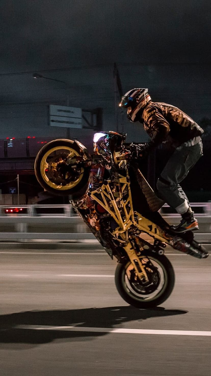 Motorcyclist performs a high wheelie on a yellow sports bike against blurred city lights at night on a dark street.