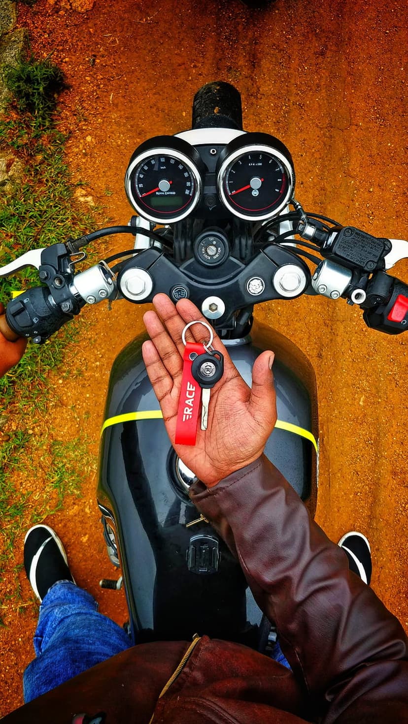 Biker in a leather jacket holds a red race tag key over a black motorcycle fuel tank with a yellow stripe.