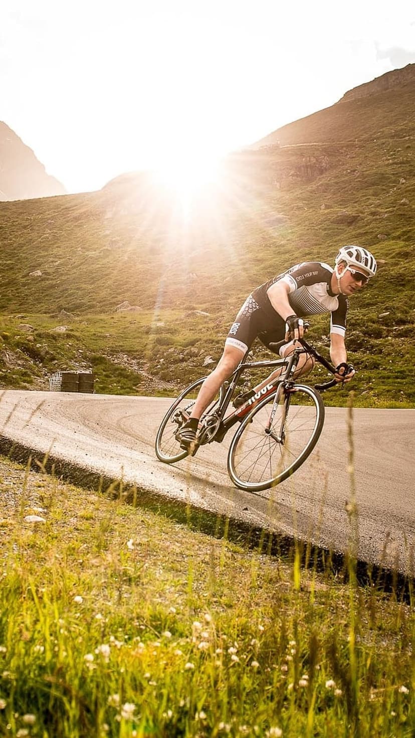 Male cyclist in racing gear leaning into a turn on a sunny mountain road with bright lens flares and grass.