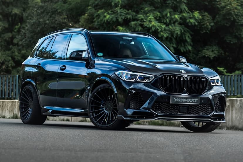 Black Manhart BMW X5 M luxury SUV with custom wheels on a road against a backdrop of green trees.