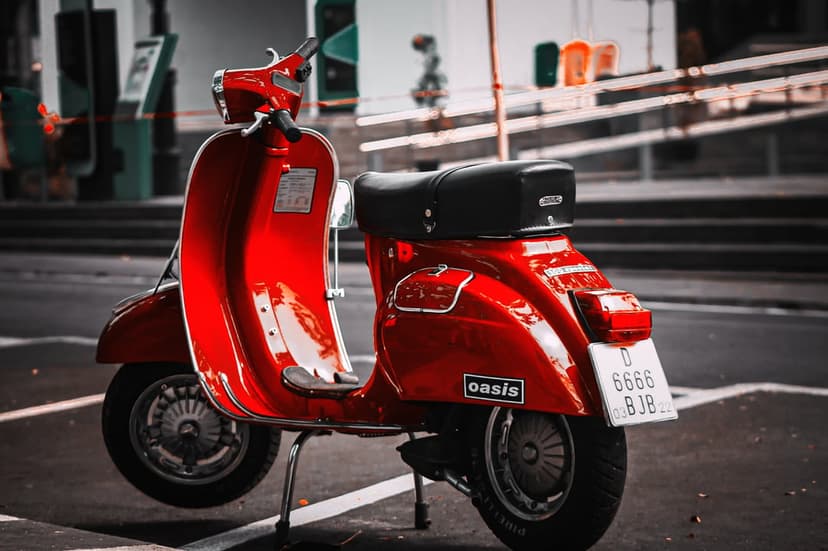 Red vintage scooter with chrome details and a black seat parked on a sunlit urban street in portrait view.