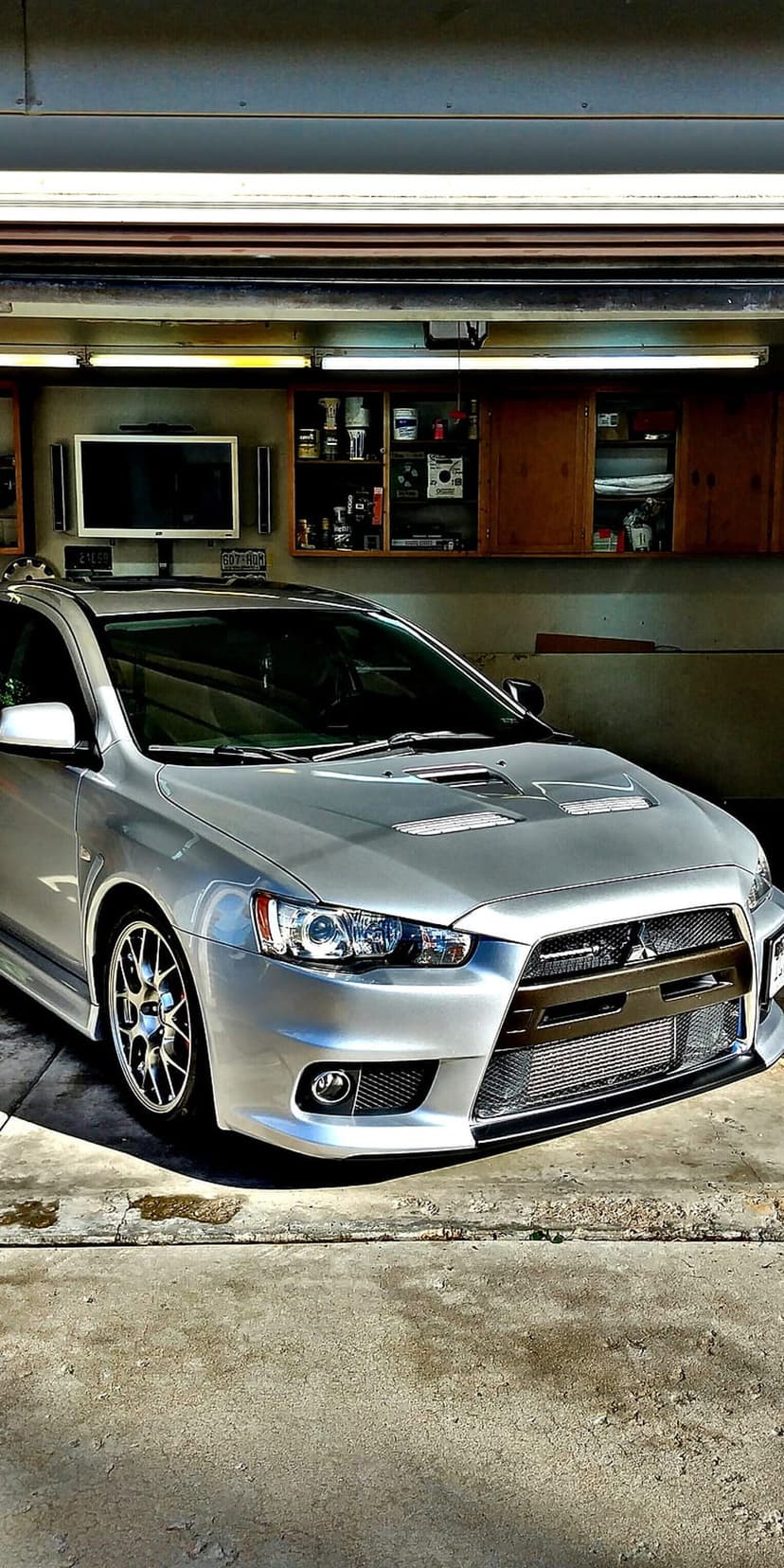 Silver Mitsubishi Lancer Evo X parked in a bright garage featuring hood vents and dark aftermarket wheels.