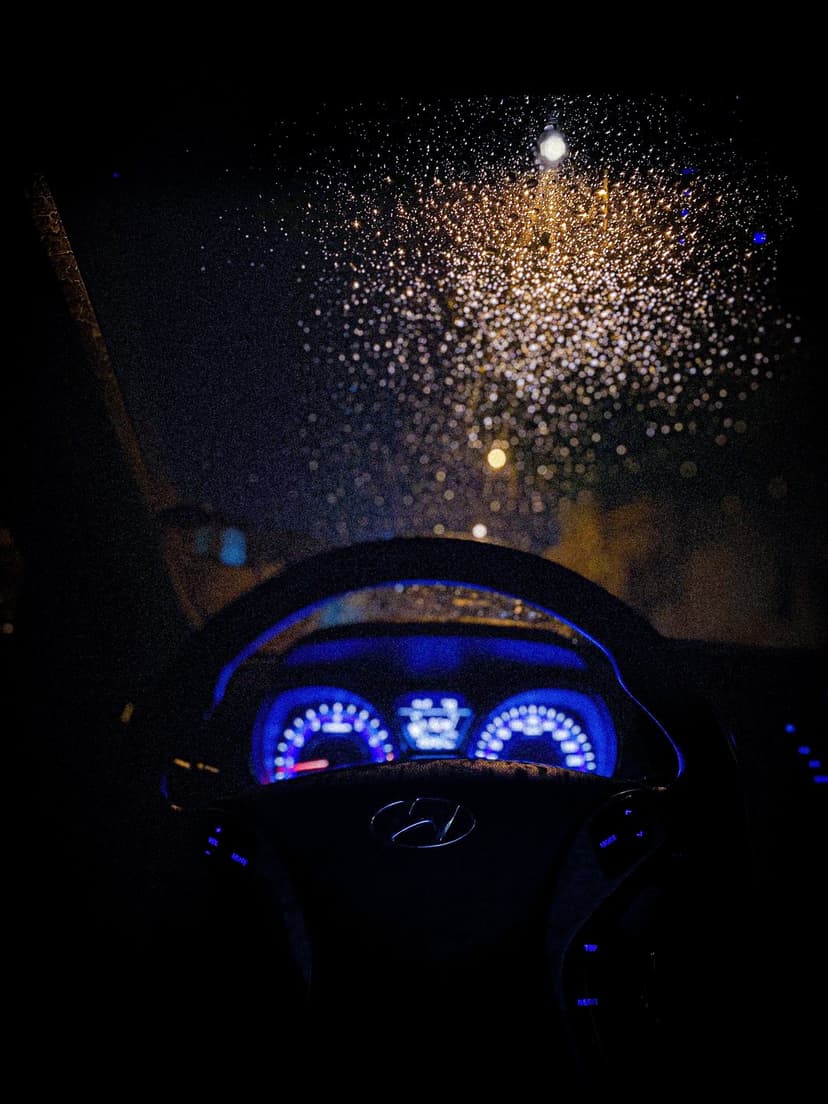 Rain-streaked windshield reveals blurred urban lights and a glowing dashboard during a night car ride.
