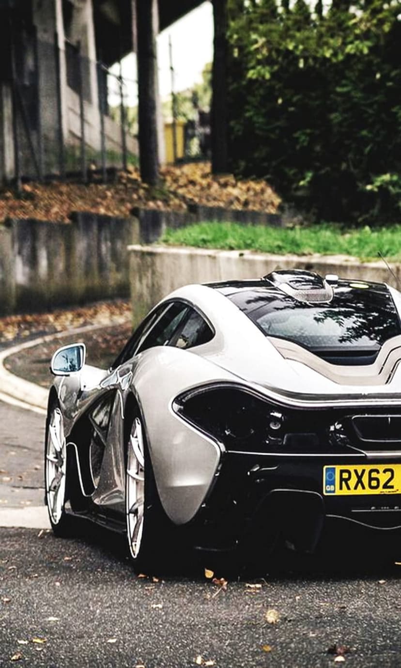 McLaren P1 rear profile on wet asphalt with glowing taillights, yellow license plate, and autumn leaves.