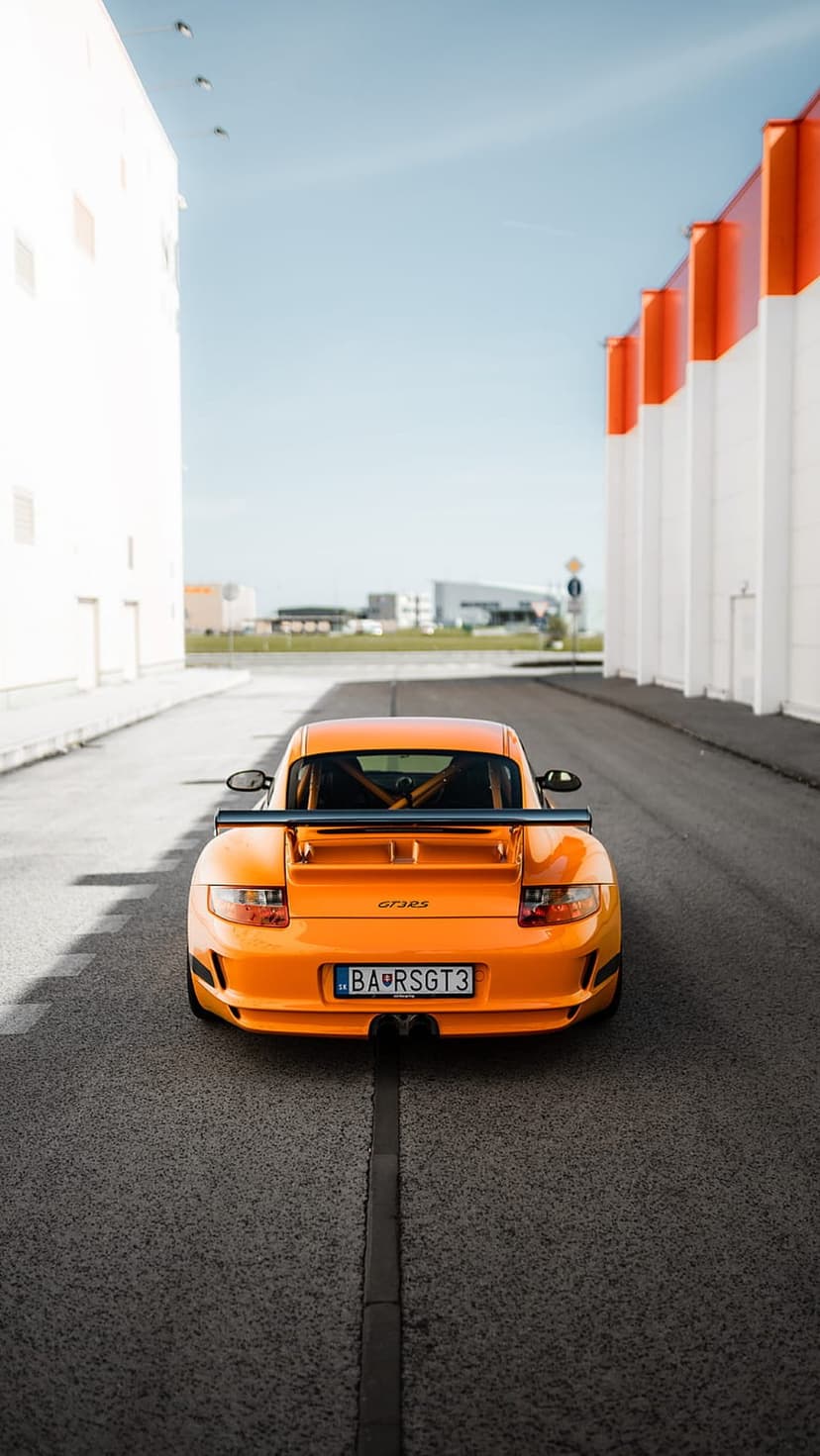 Porsche 911 GT3 RS in orange shows its massive rear wing and sleek tail lights on an asphalt industrial road.