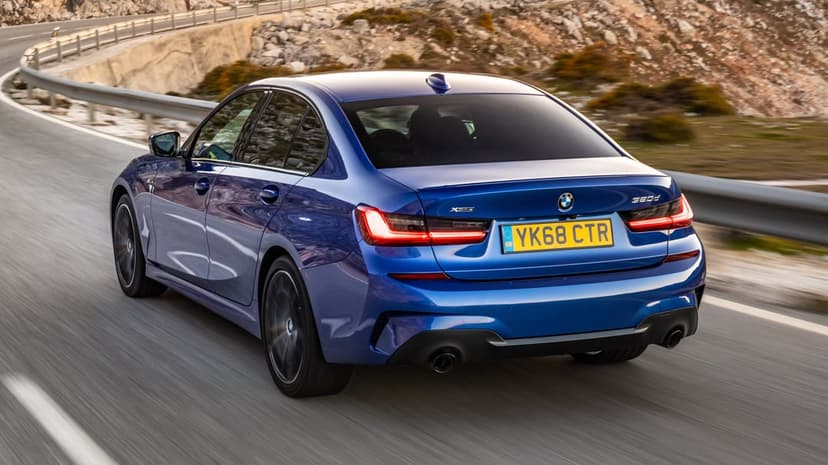 Rear view of a blue BMW sedan driving on a winding mountain road with motion blur and rocky terrain background.