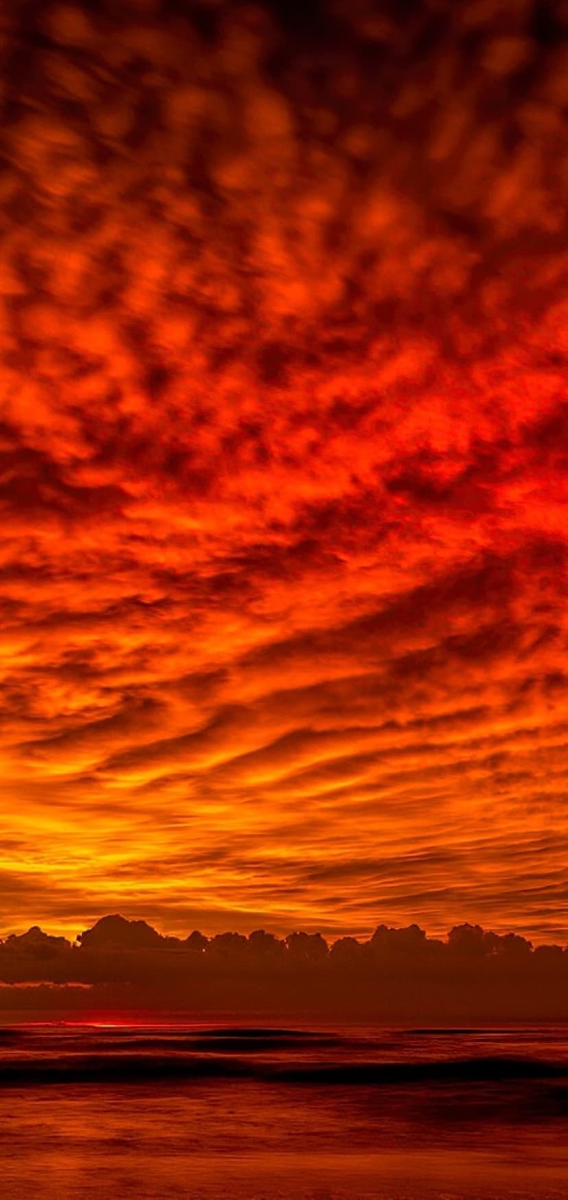 Fiery red and orange clouds stretch horizontally across a dark ocean horizon during a vivid coastal sunset.