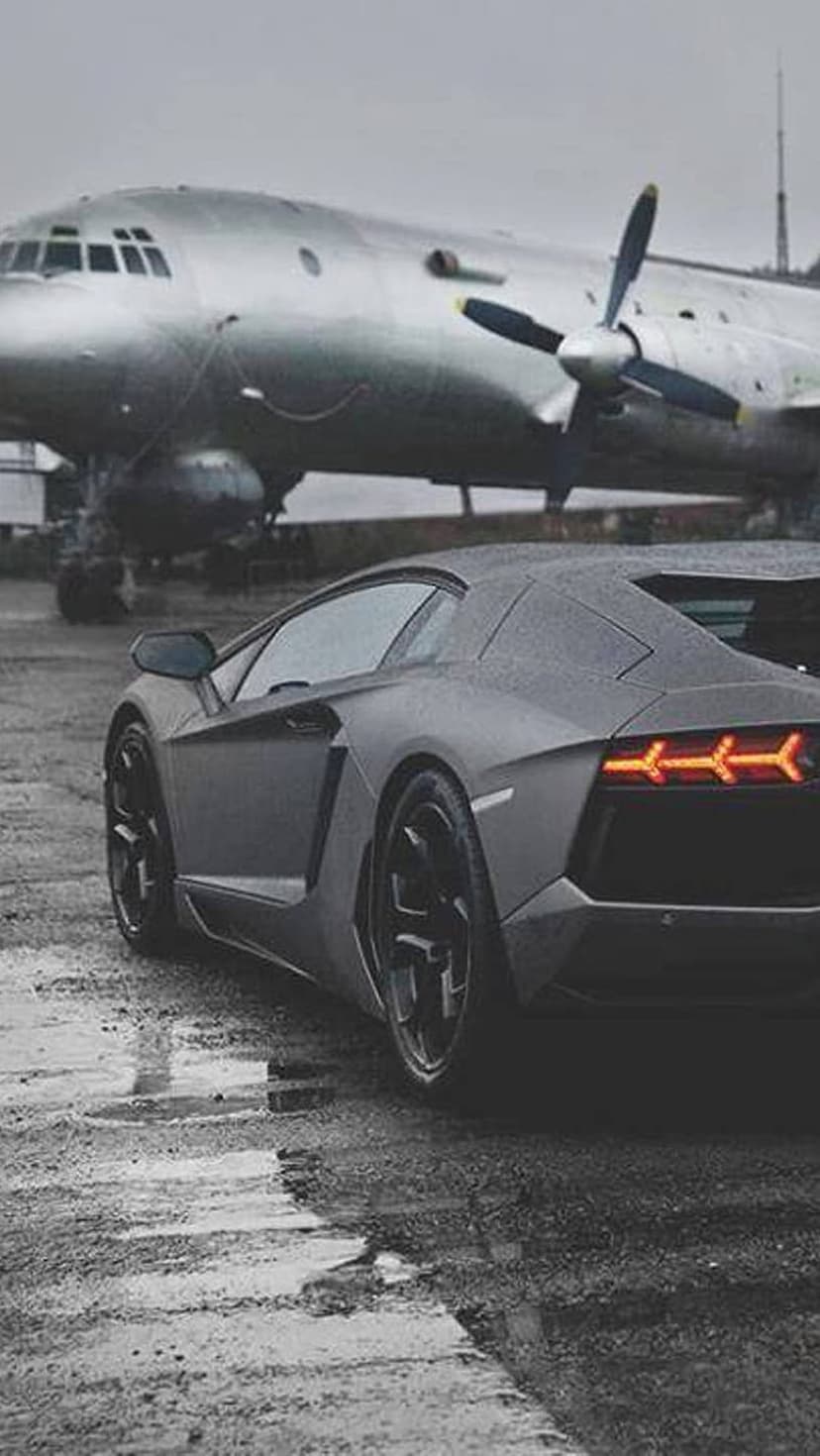 Charcoal gray supercar parked beside a large silver vintage propeller plane on a rainy, reflective runway.