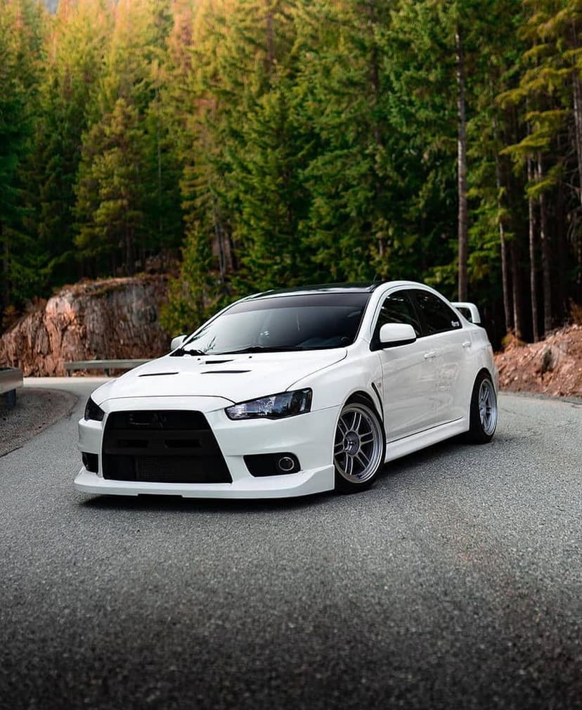 White Mitsubishi Lancer Evolution driving on a winding asphalt road through a dense forest of evergreen trees.