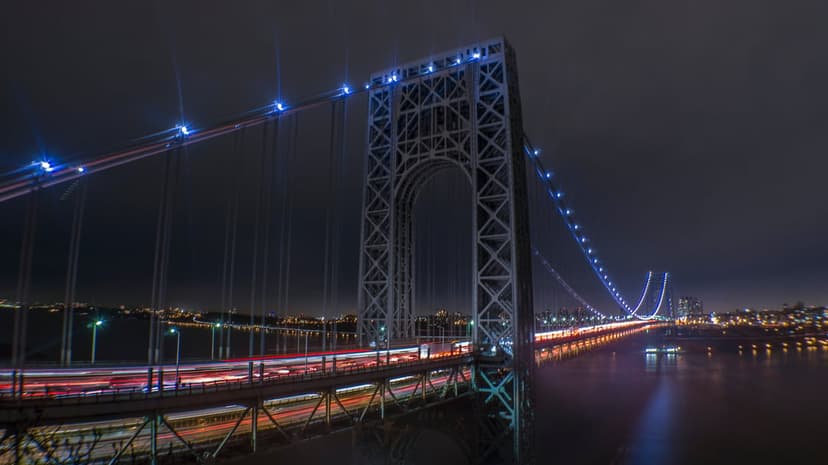 George Washington Bridge at night with blue cable lights, red traffic streaks, and reflections on the Hudson River.