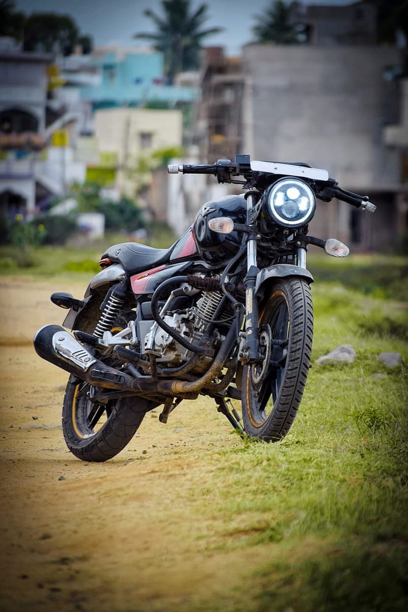 Custom black motorcycle with a lit circular LED headlight parked on a gravel road against blurred buildings.