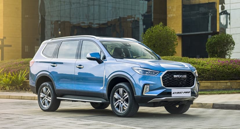 Metallic blue Maxus D90 Pro SUV parked on a stone plaza in front of a glass building at sunset.